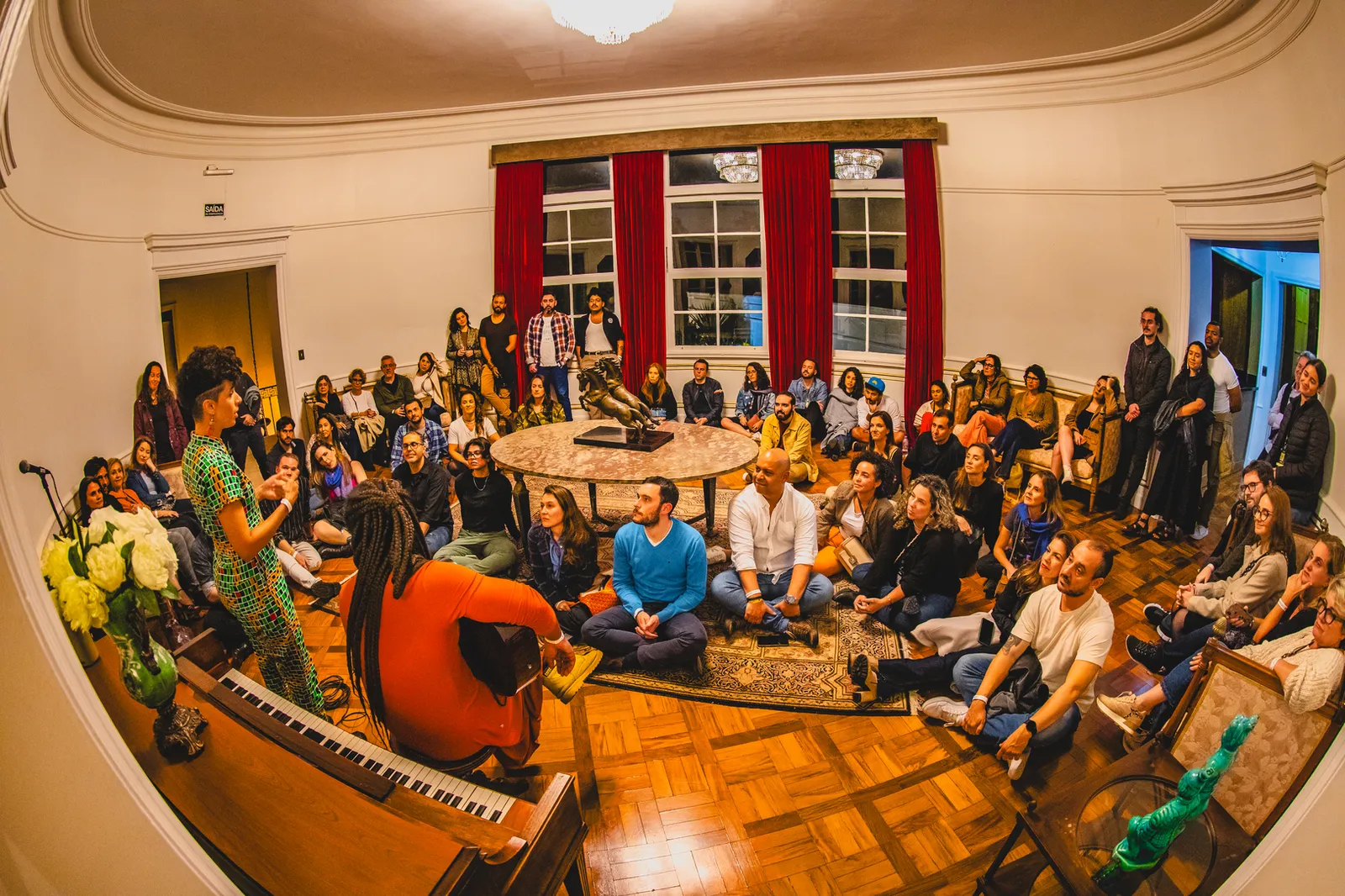 Artista conduz uma conversa ou apresentação para o público reunido em uma sala histórica do Nacional Club. A imagem representa os espaços dedicados a encontros, palestras e experiências culturais da Jazz Mansion.