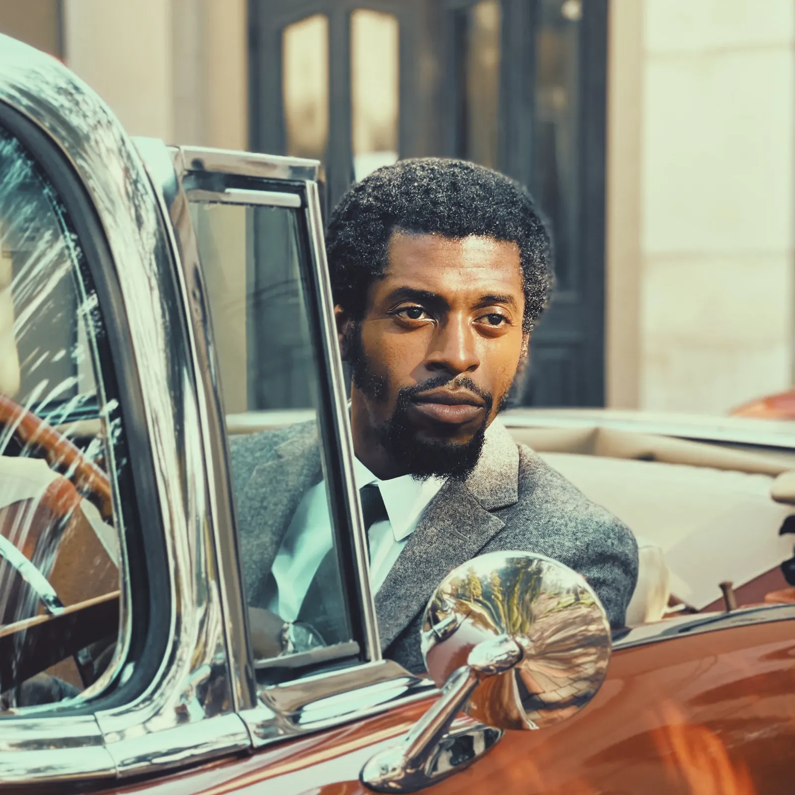 Artista veste terno cinza e observa o ambiente sentado em um carro conversível clássico. A imagem apresenta um dos nomes da programação artística da Jazz Mansion.